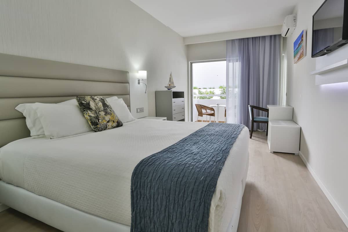 A hotel room with a large bed, a desk, a chair, a TV, and a balcony with a view.