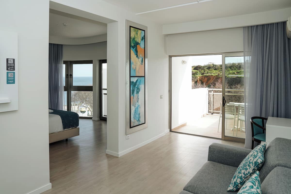 A modern living room with a grey sofa, a large abstract painting, and a balcony with a view of the sea.