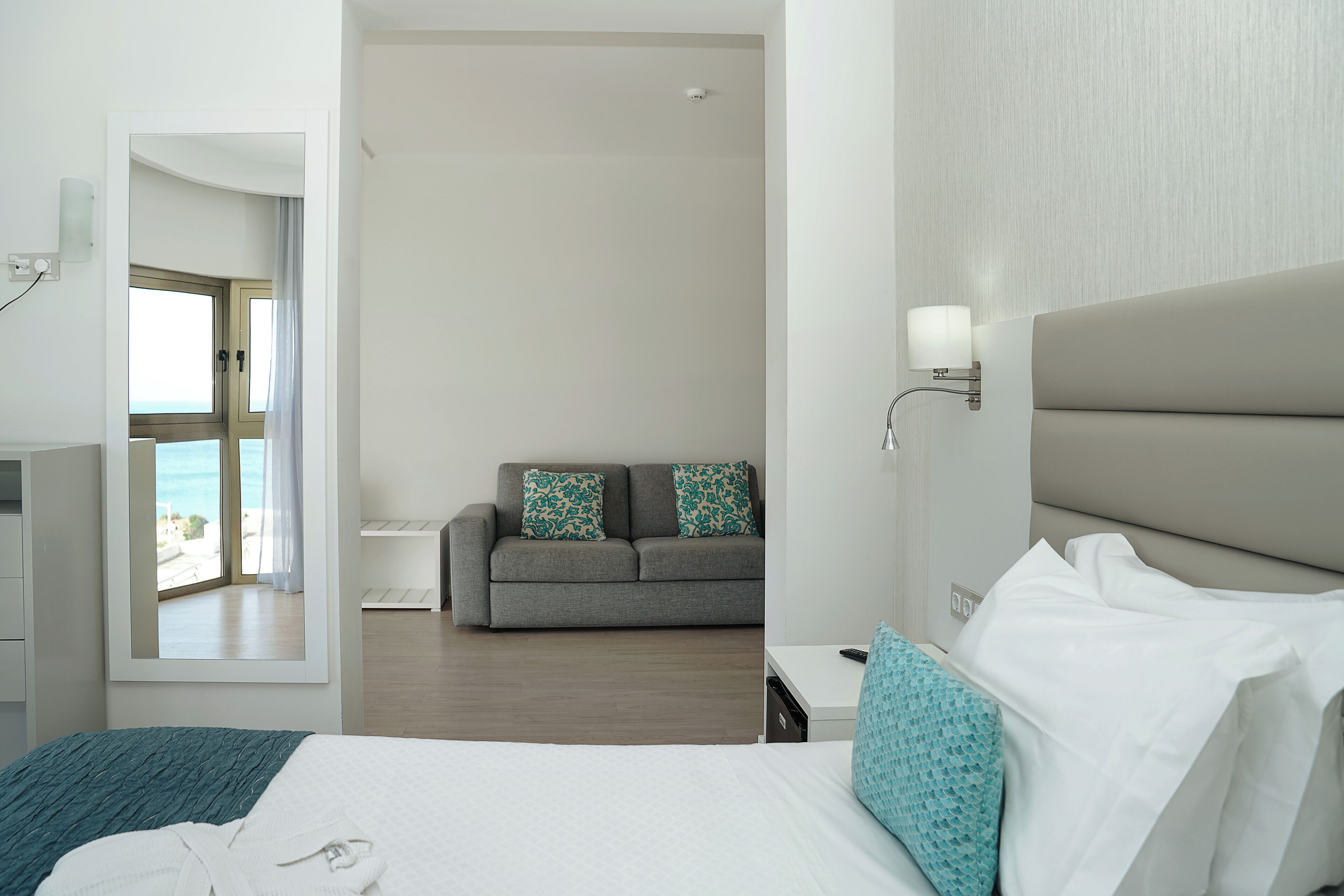 Une chambre d’hôtel avec un lit, un canapé, une table de chevet et une vue sur la mer, visible par la porte.
