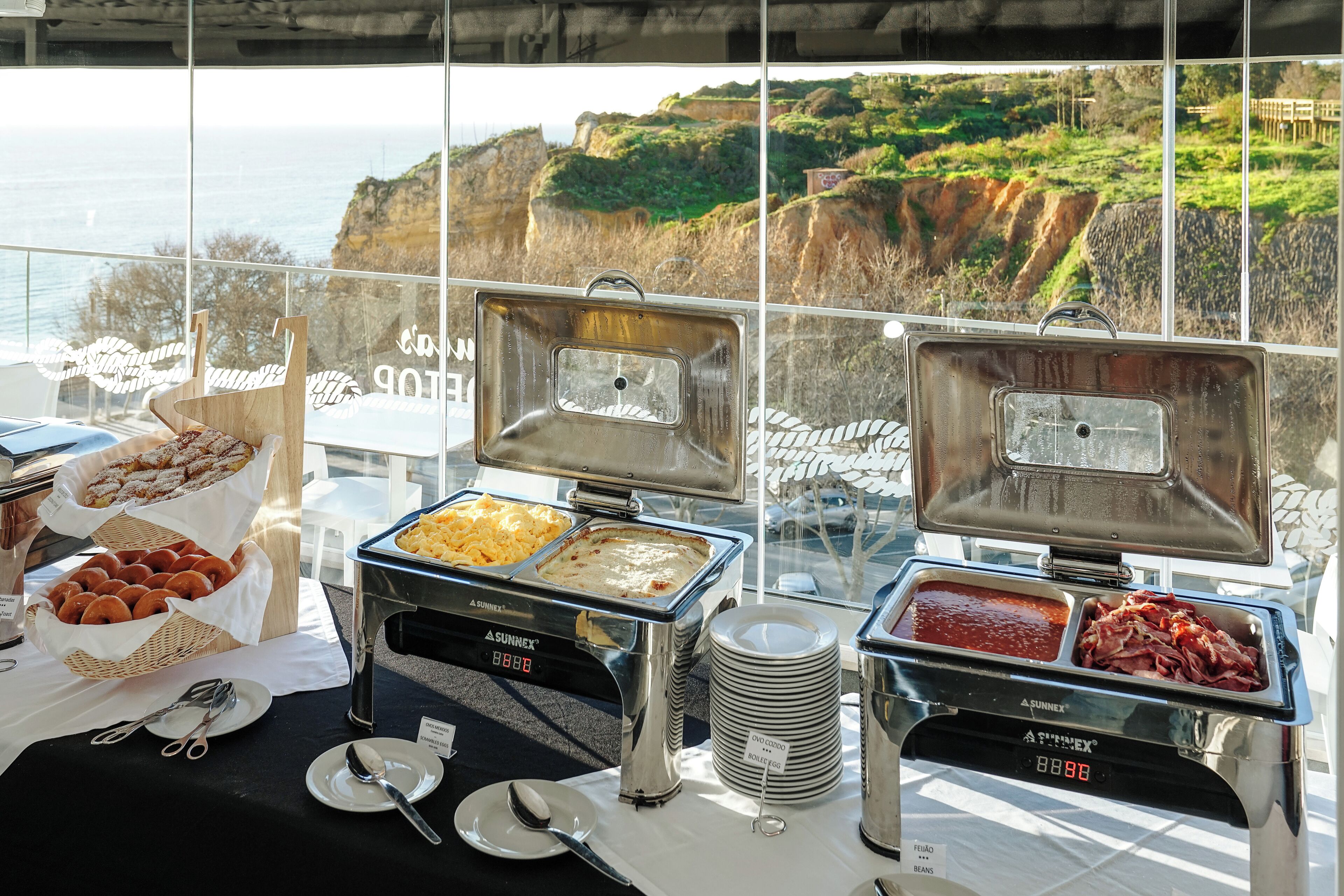 Una mesa de buffet con diversos alimentos y utensilios, con un pintoresco paisaje costero de fondo.