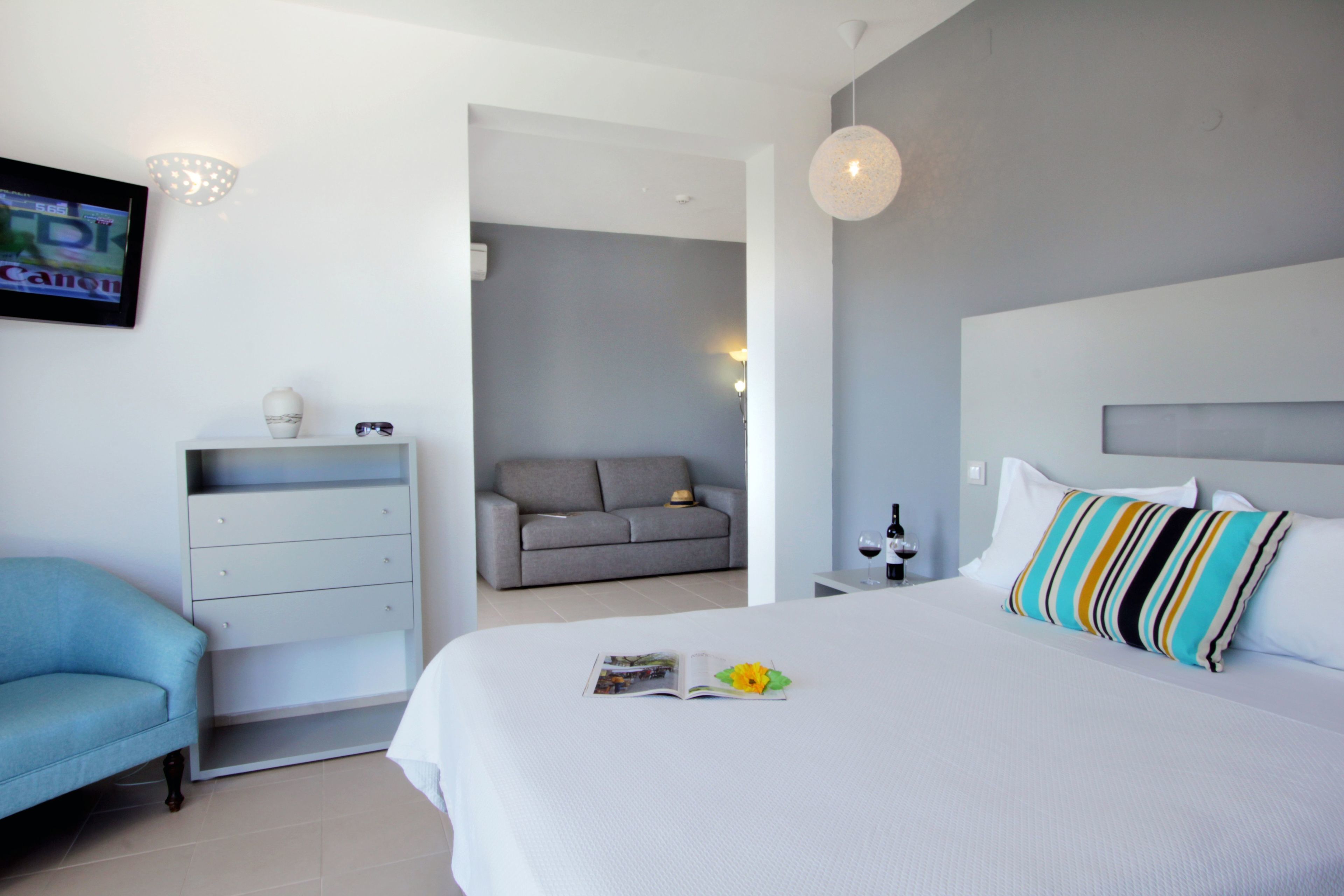 A modern hotel room with a large bed, a grey sofa, a small table with a wine bottle, and a TV on the wall.