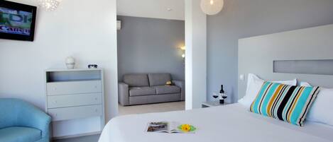 A modern hotel room with a large bed, a grey sofa, a small table with a wine bottle, and a TV on the wall.