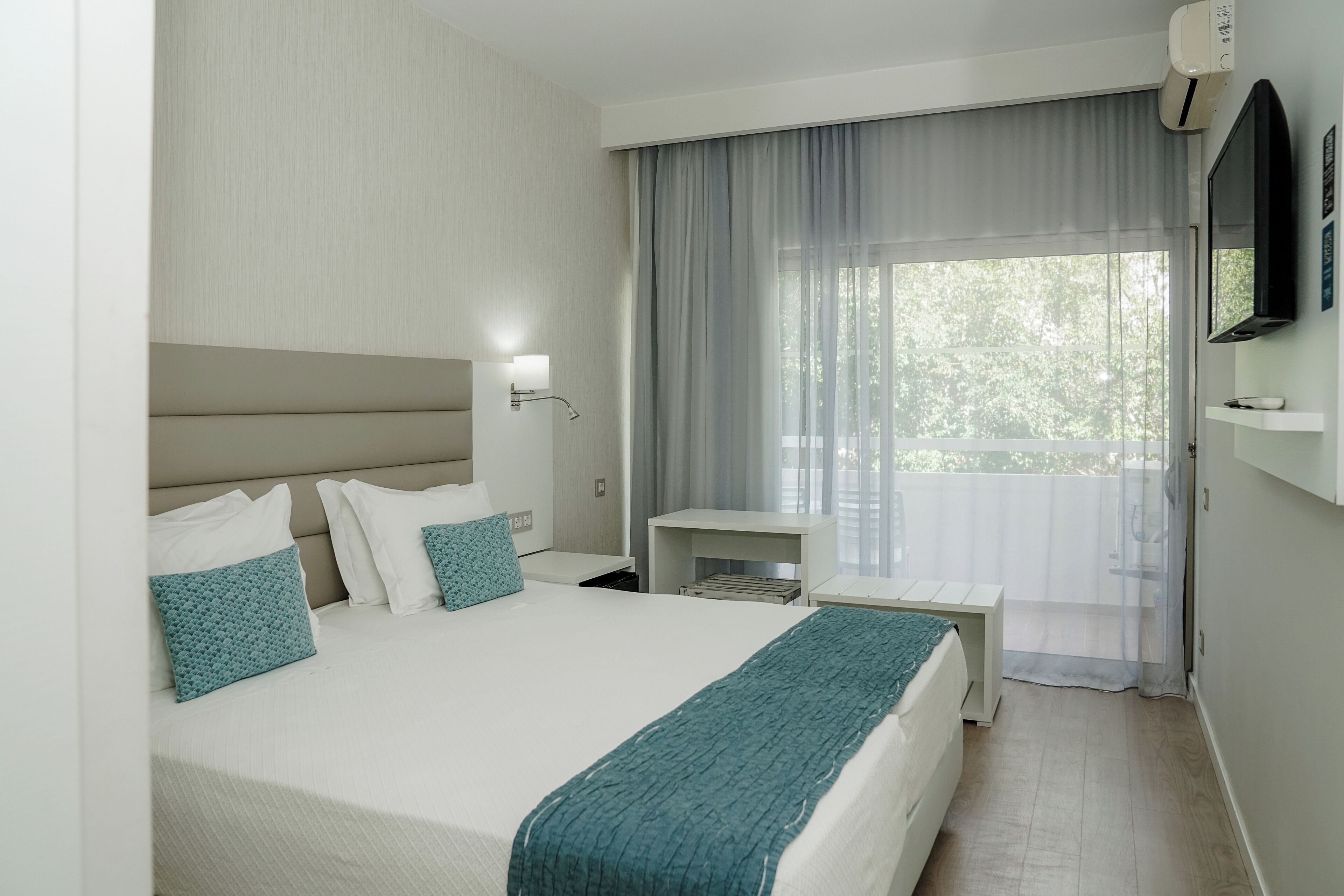 Ein Hotelzimmer mit Bett, Nachttisch, an der Wand befestigtem Fernseher und einem Fenster mit Vorhängen.