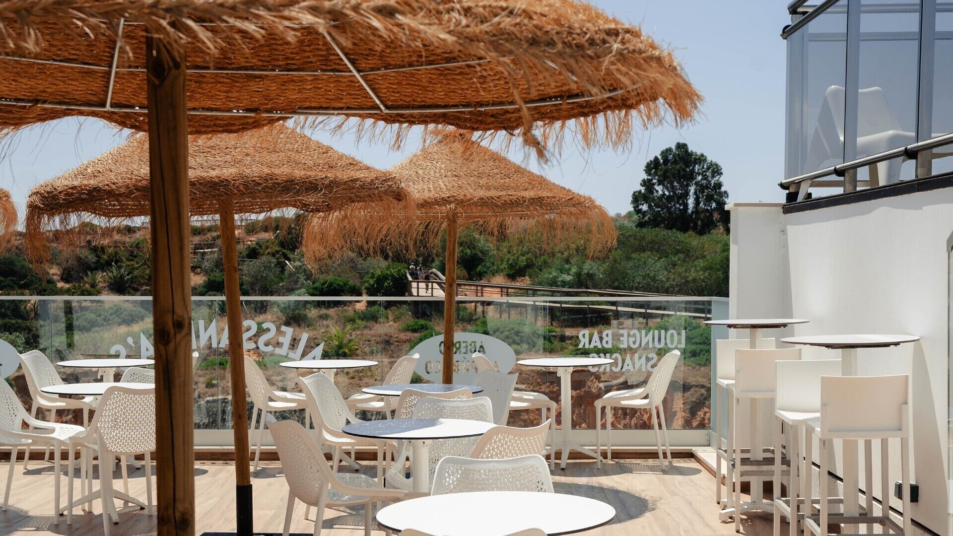 An outdoor seating area with white plastic chairs and round tables, covered by thatched umbrellas, overlooking a scenic landscape.