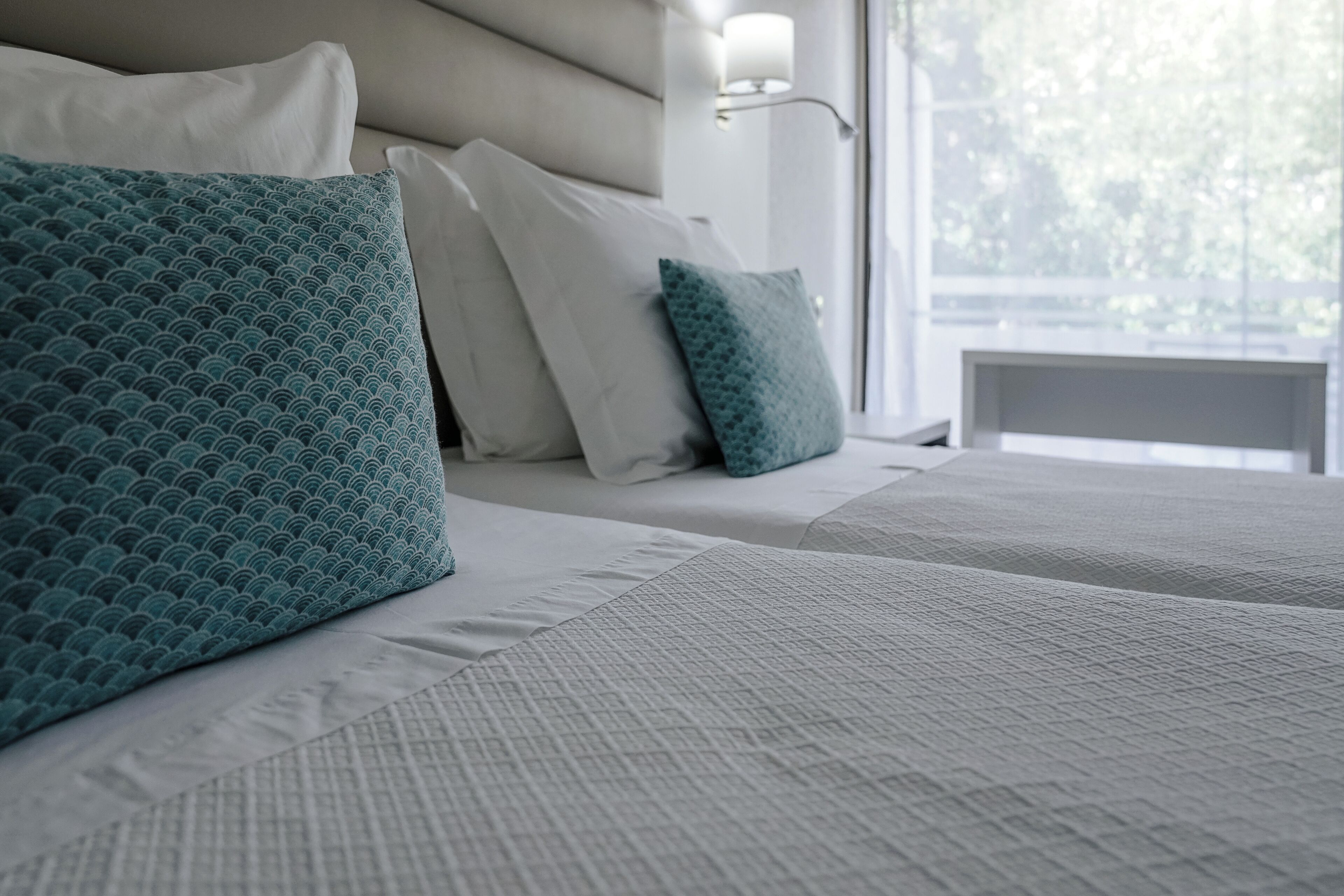 A neatly made bed with white and teal pillows, a textured white bedspread, and a bedside lamp.