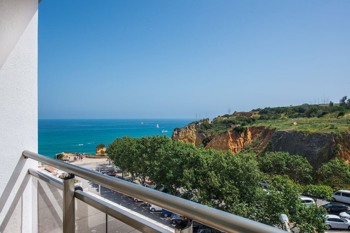 A balcony with a view of the sea, a parking area, and a cliffside with greenery.