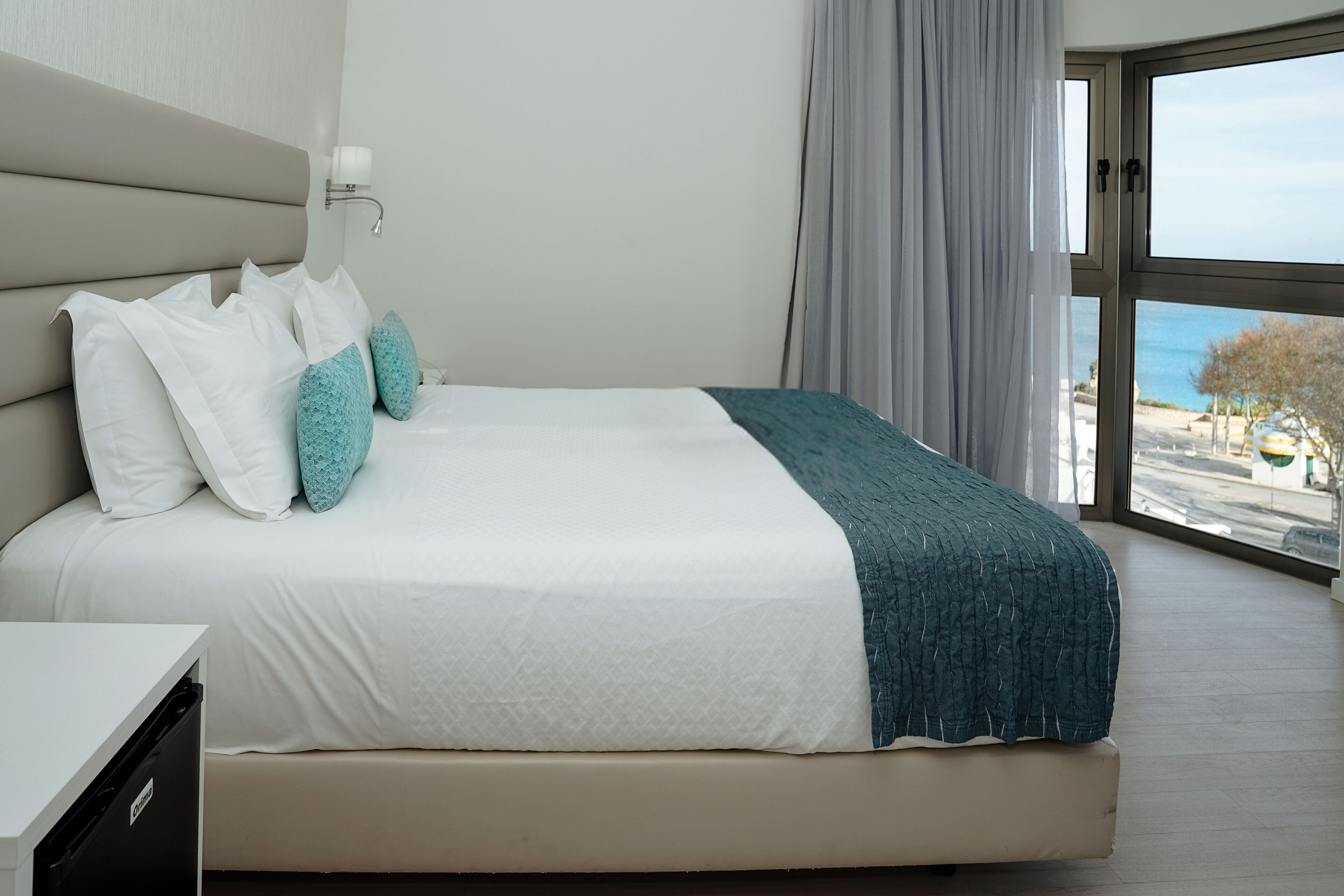 A modern hotel room with a large bed, a nightstand, and a view of the sea through a large window.