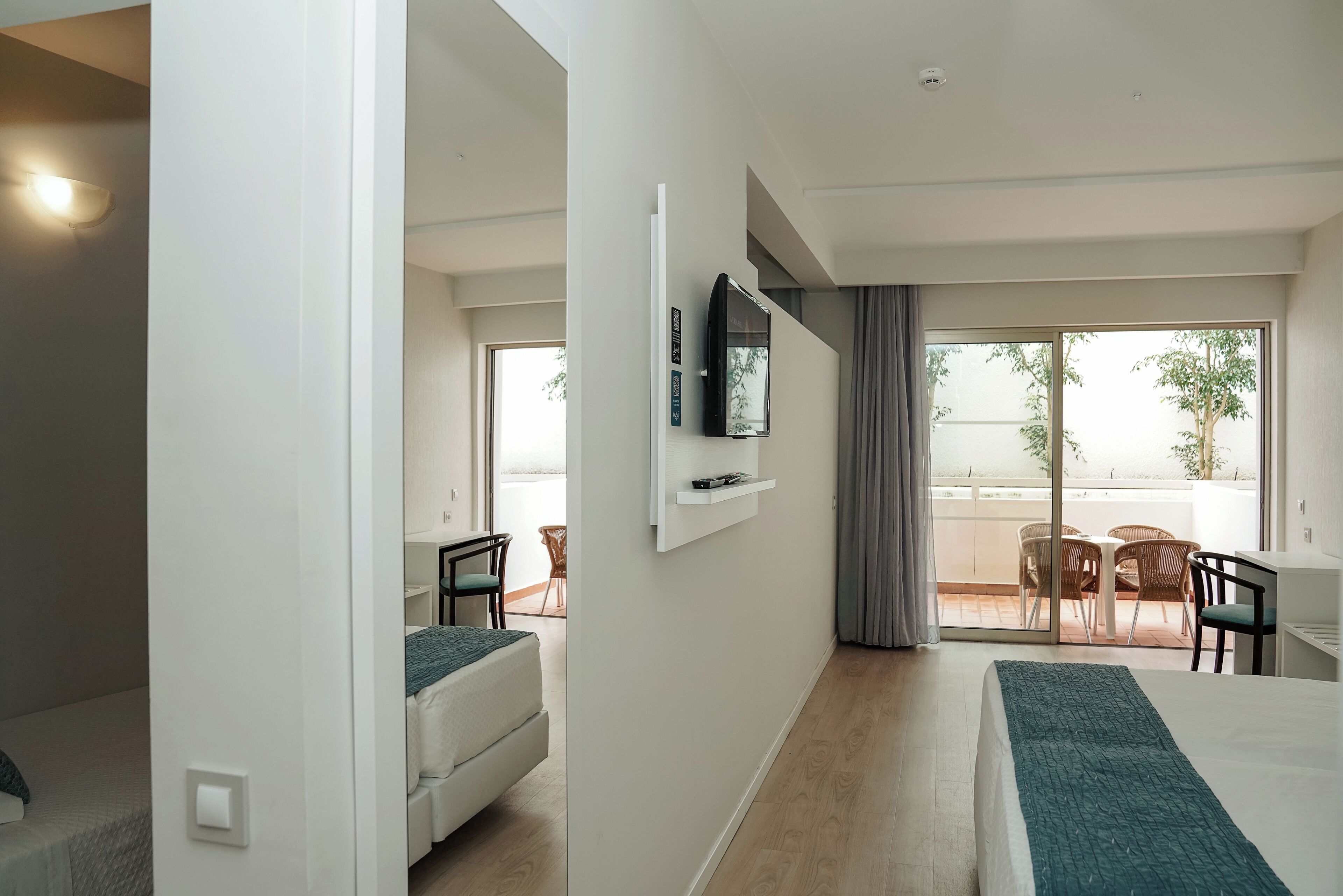 Quarto de hotel com cama, escrivaninha, cadeira, televisão na parede e varanda com mesa e cadeiras.