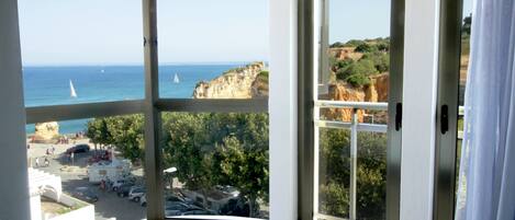 A hotel room with a large bay window offering a view of the sea, cliffs, and a parking area.