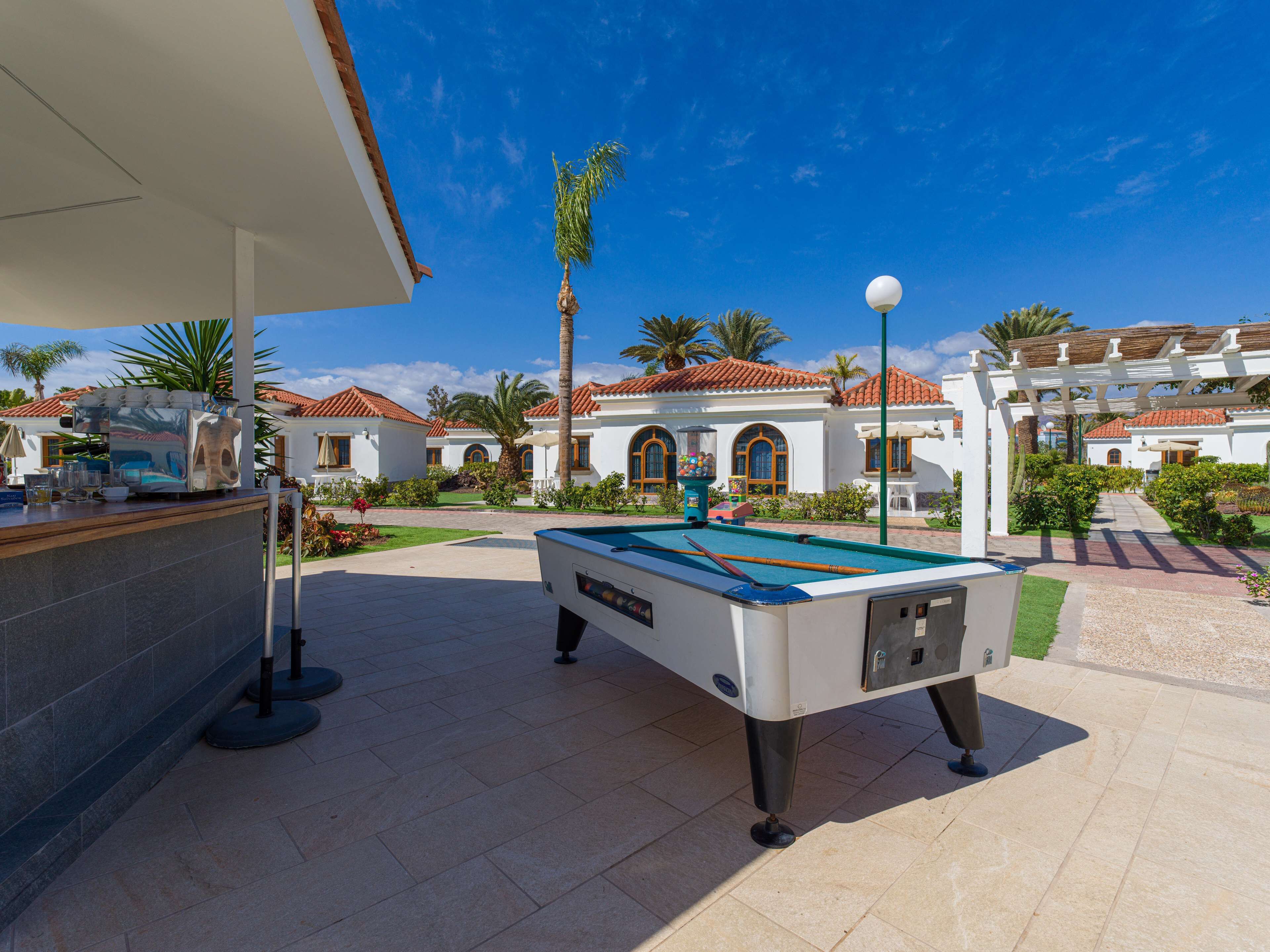 A pool table in an outdoor area with a patio, a bar, and a building with a tiled roof.