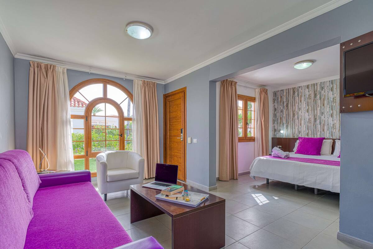 Une chambre d’hôtel avec un canapé violet, un bureau en bois avec des livres, un lit avec une literie blanche et rose, et une fenêtre avec des rideaux.