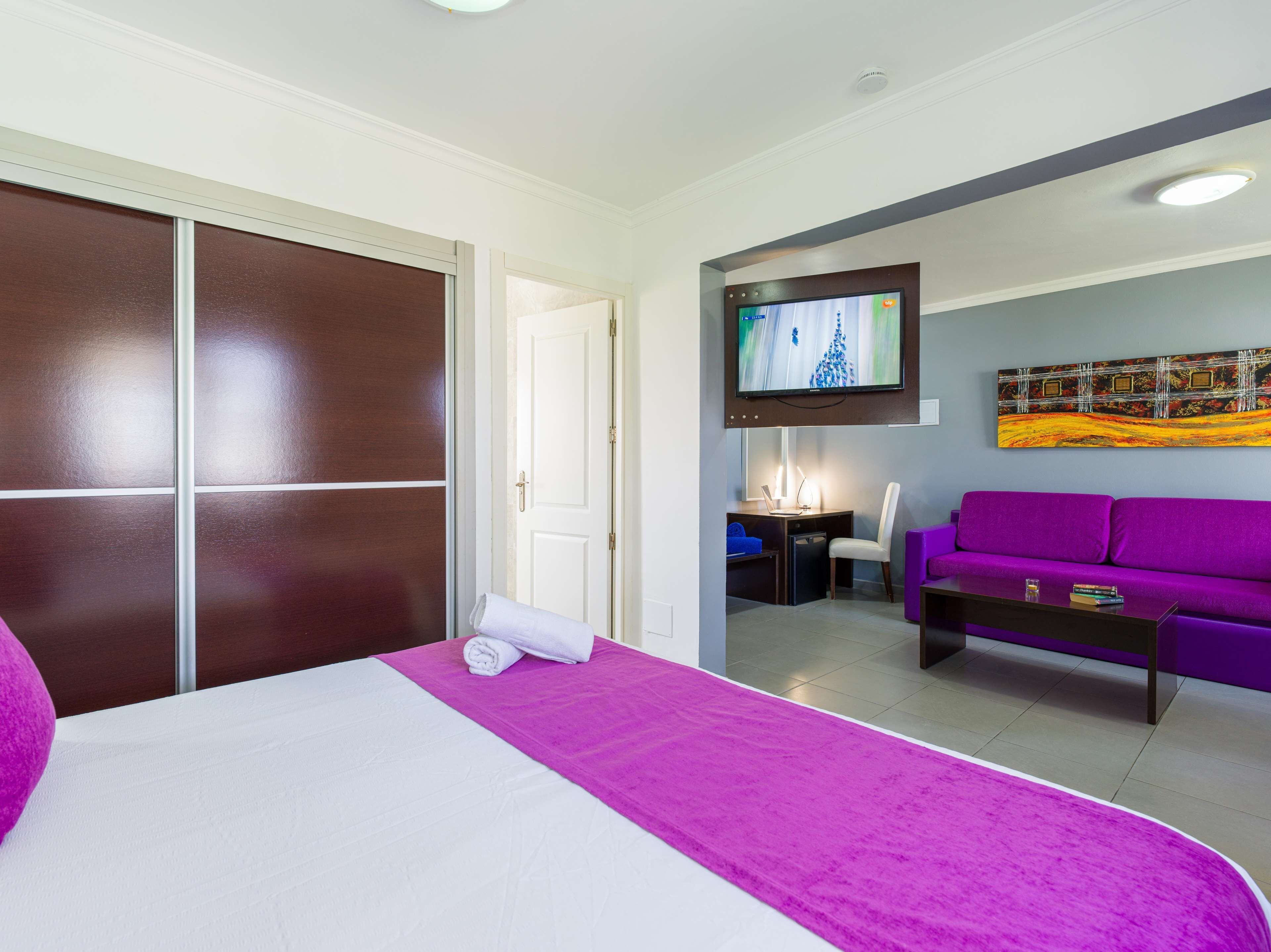 Ein Hotelzimmer mit einem Bett, einem Schreibtisch mit Stuhl, einem violetten Sofa, einem flachbildfernseher an der Wand und einem Bild über dem Fernseher.