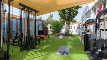 Une salle de sport équipée de matériel d’exercice, un espace abrité avec un auvent, et un petit jardin avec un arbre et des plantes.