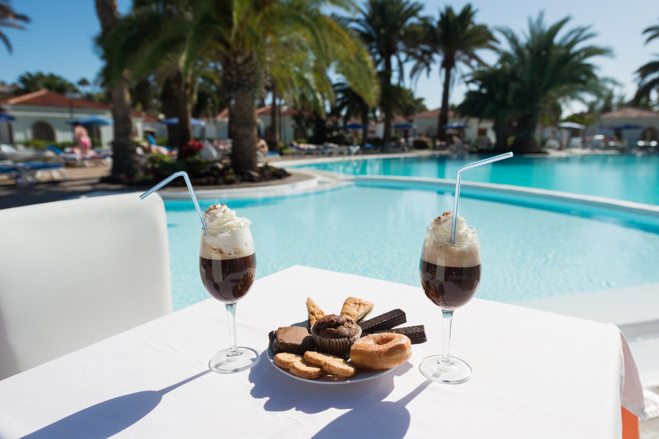 A poolside table with a plate of pastries and two glasses of a dark beverage, with a pool and palm trees in the background.