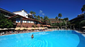 Una amplia piscina con tumbonas y sombrillas, rodeada por un hotel de varios pisos y palmeras.