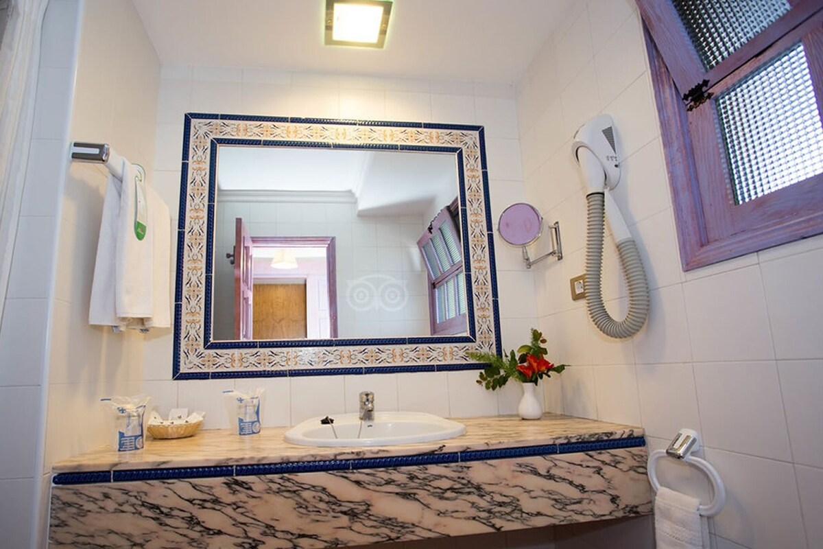 Un baño con lavamanos de mármol, un espejo grande con marco decorativo, un cabezal de ducha de mano y un perchero.