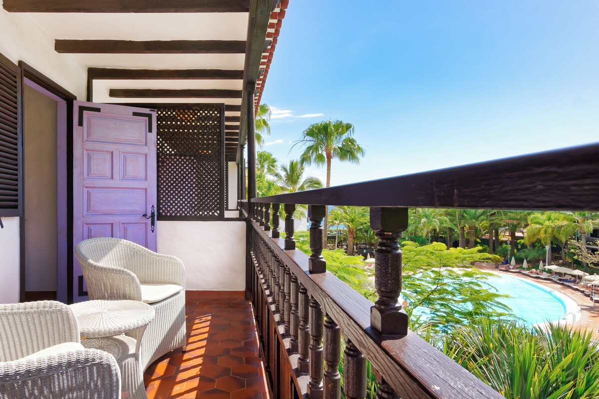 Un balcón con mobiliario de mimbre, una puerta morada y vistas a una piscina y palmeras.