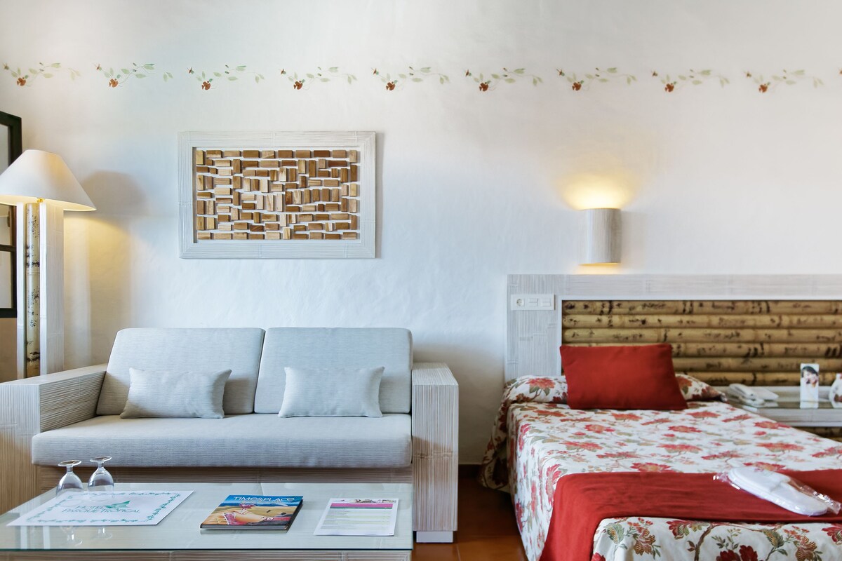 Habitación de hotel con sofá, cama con ropa de cama floral, un adorno en la pared y una mesita de noche con lámpara.