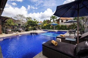 Una piscina con sillas de descanso y sombrillas, rodeada de vegetación y edificios.