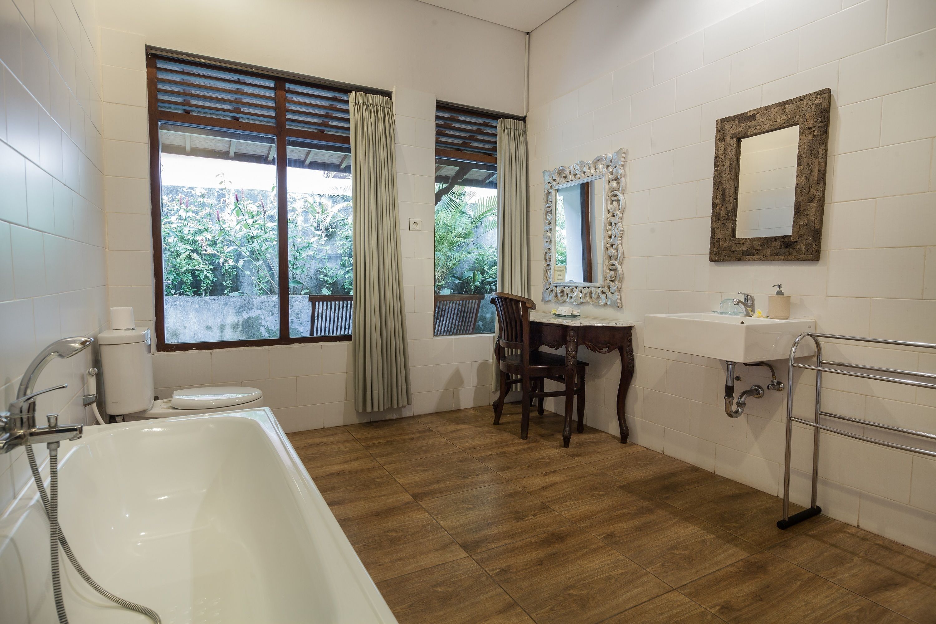Un baño con bañera, lavamanos y espejo. Hay dos ventanas con cortinas, y una silla está colocada cerca del lavamanos.