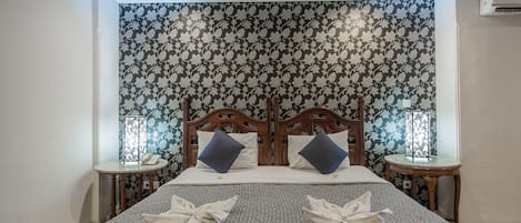 Ein ordentlich gemachtes Bett mit einem gemusterten Bettbezug, zwei hölzerne Nachttische mit Lampen und eine blumige Tapete im Hintergrund.