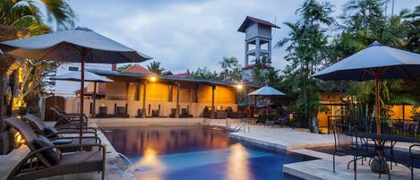 A swimming pool area with lounge chairs and umbrellas, a building with a red roof, and palm trees in the background.