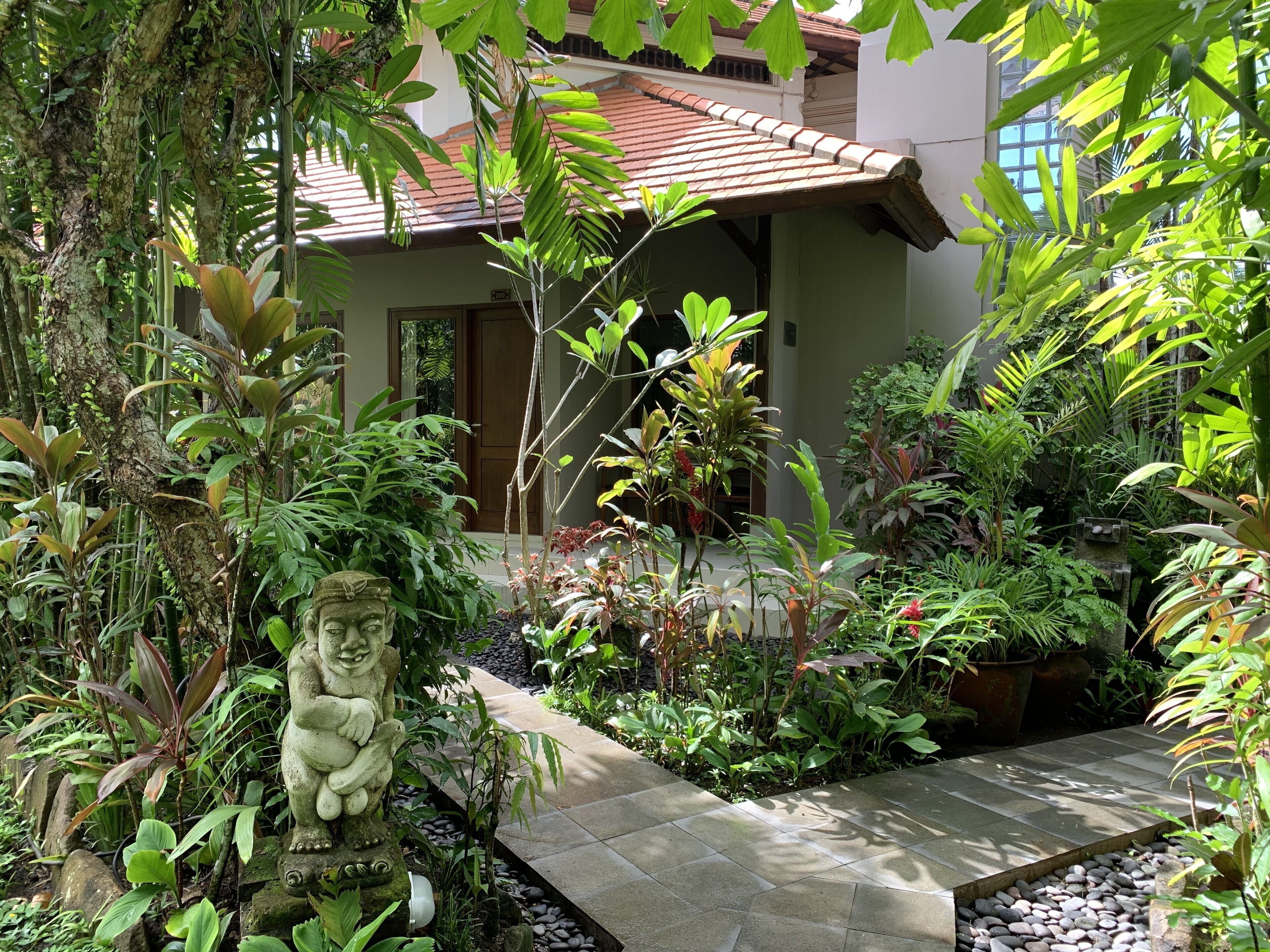 A lush garden with a stone statue, a tiled pathway, and a building with a tiled roof.