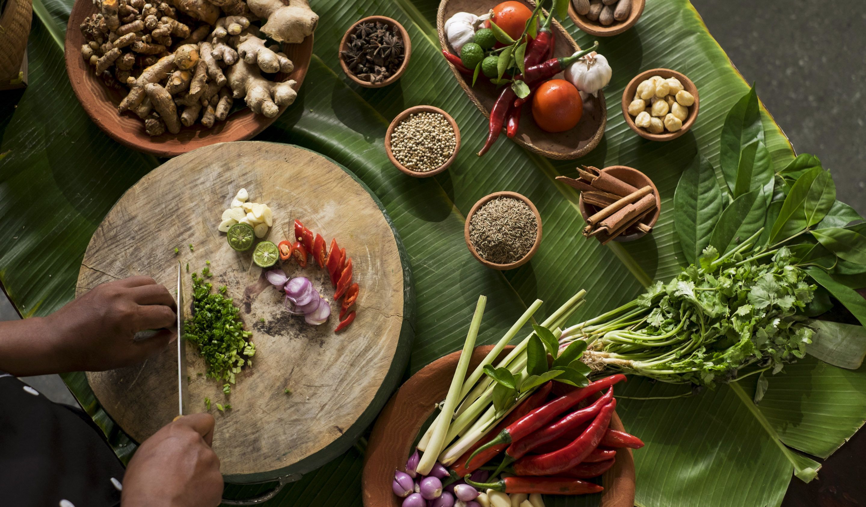 Variedad de ingredientes frescos, como jengibre, ajo, tomates, chiles y hierbas, dispuestos sobre una tabla de madera y hojas de plátano.