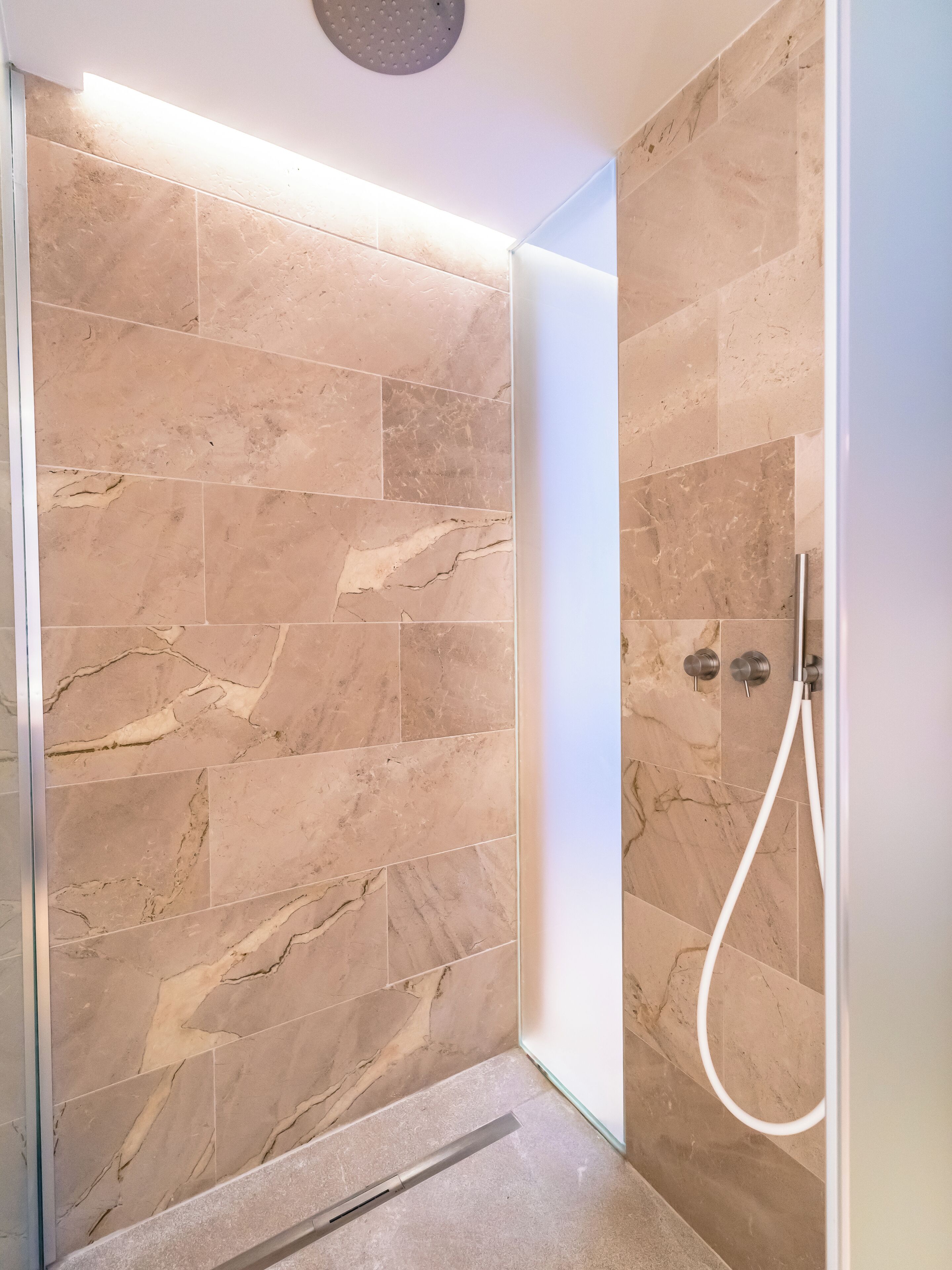 Un área de ducha moderna con baldosas de mármol color crema y puerta de vidrio.