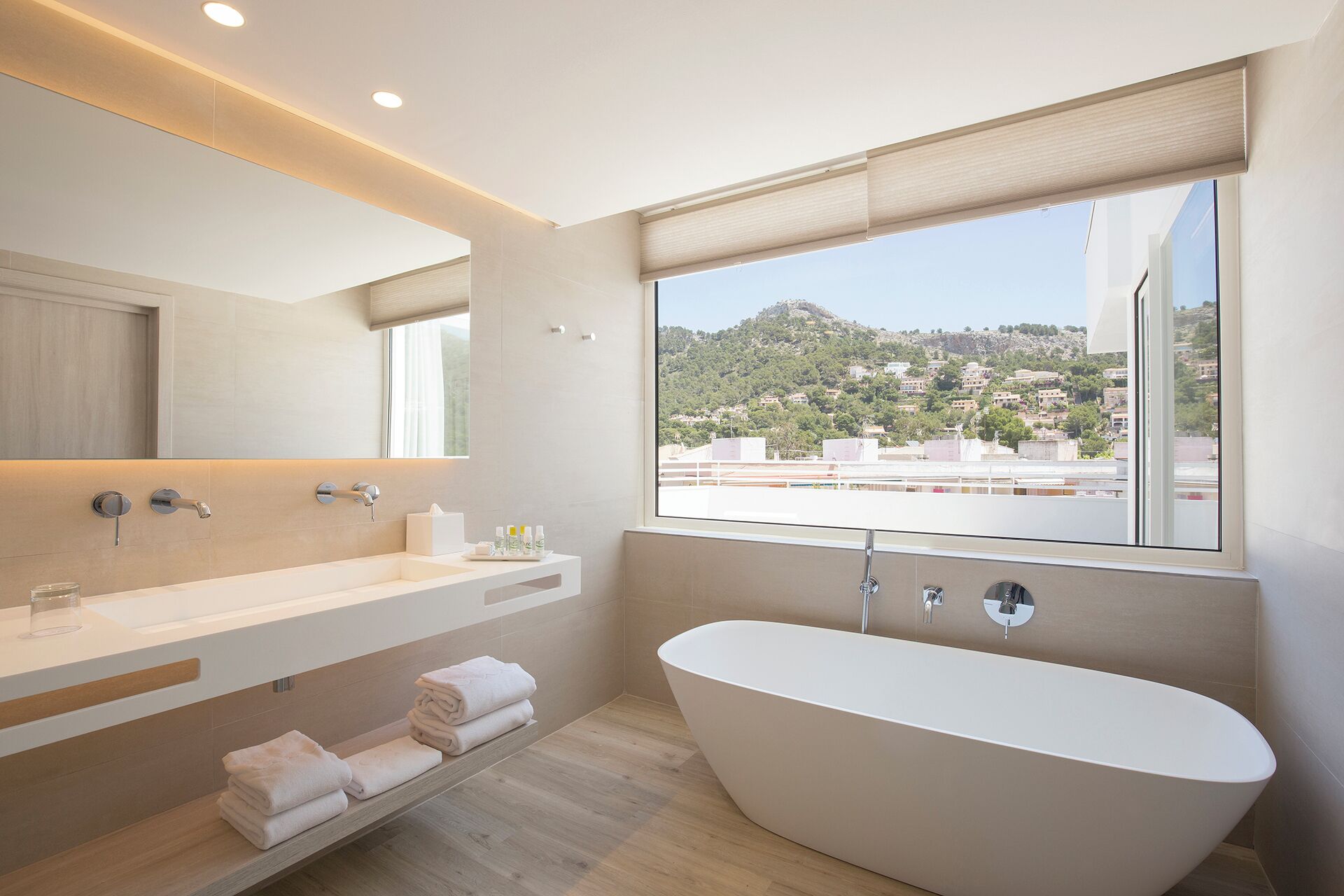 Ein modernes Badezimmer mit freistehender Badewanne, einem Waschtisch inklusive Spiegel und einem Fenster mit Blick auf eine Hügellandschaft.