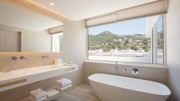 Ein modernes Badezimmer mit freistehender Badewanne, einem Waschtisch inklusive Spiegel und einem Fenster mit Blick auf eine Hügellandschaft.