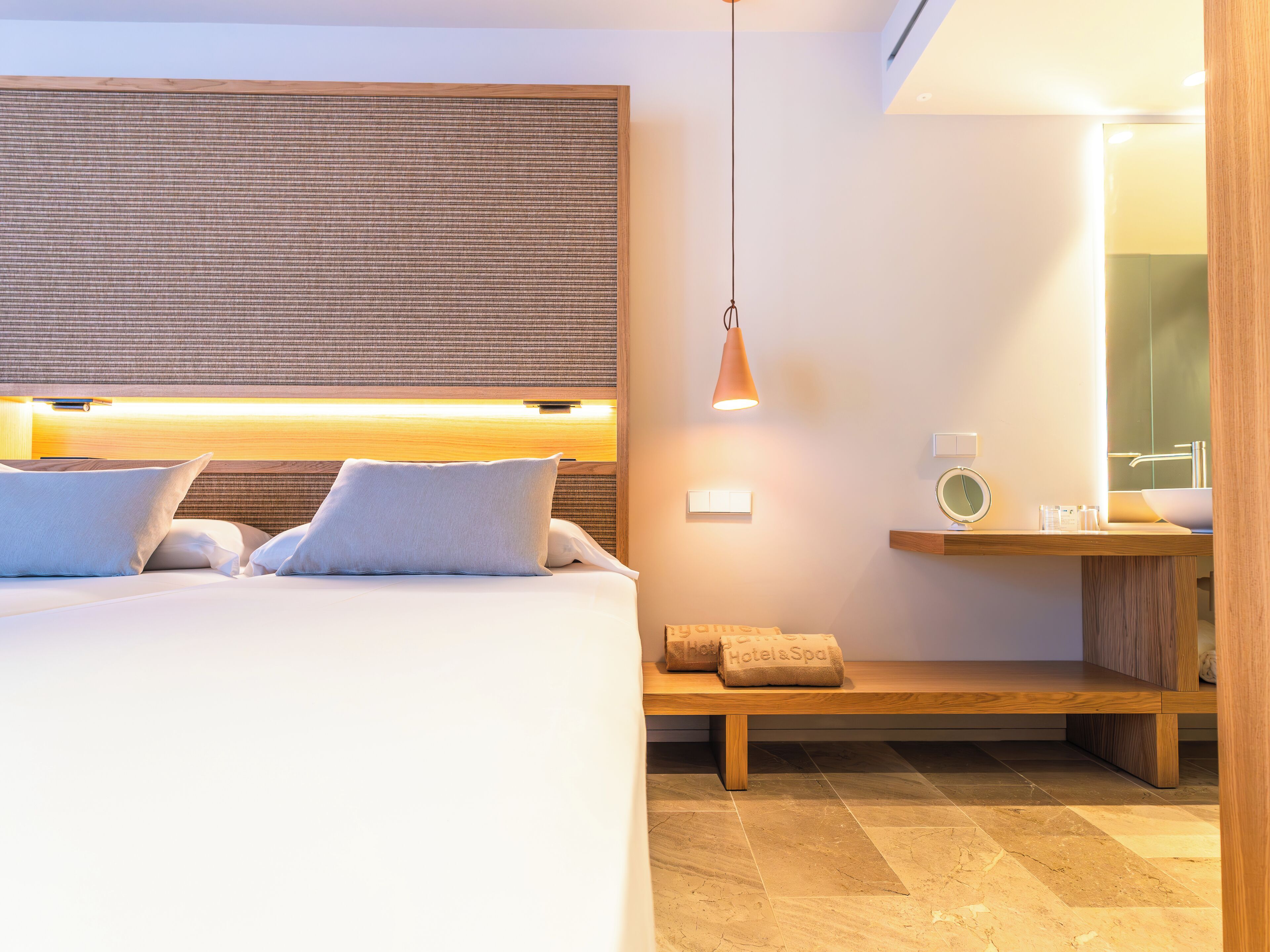 A modern hotel room with a large bed, a bedside table with a lamp, a mirror, and a bench with towels.