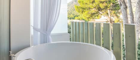 A modern bathroom with a freestanding bathtub, a shower curtain, and a view of a green landscape with trees and hills.