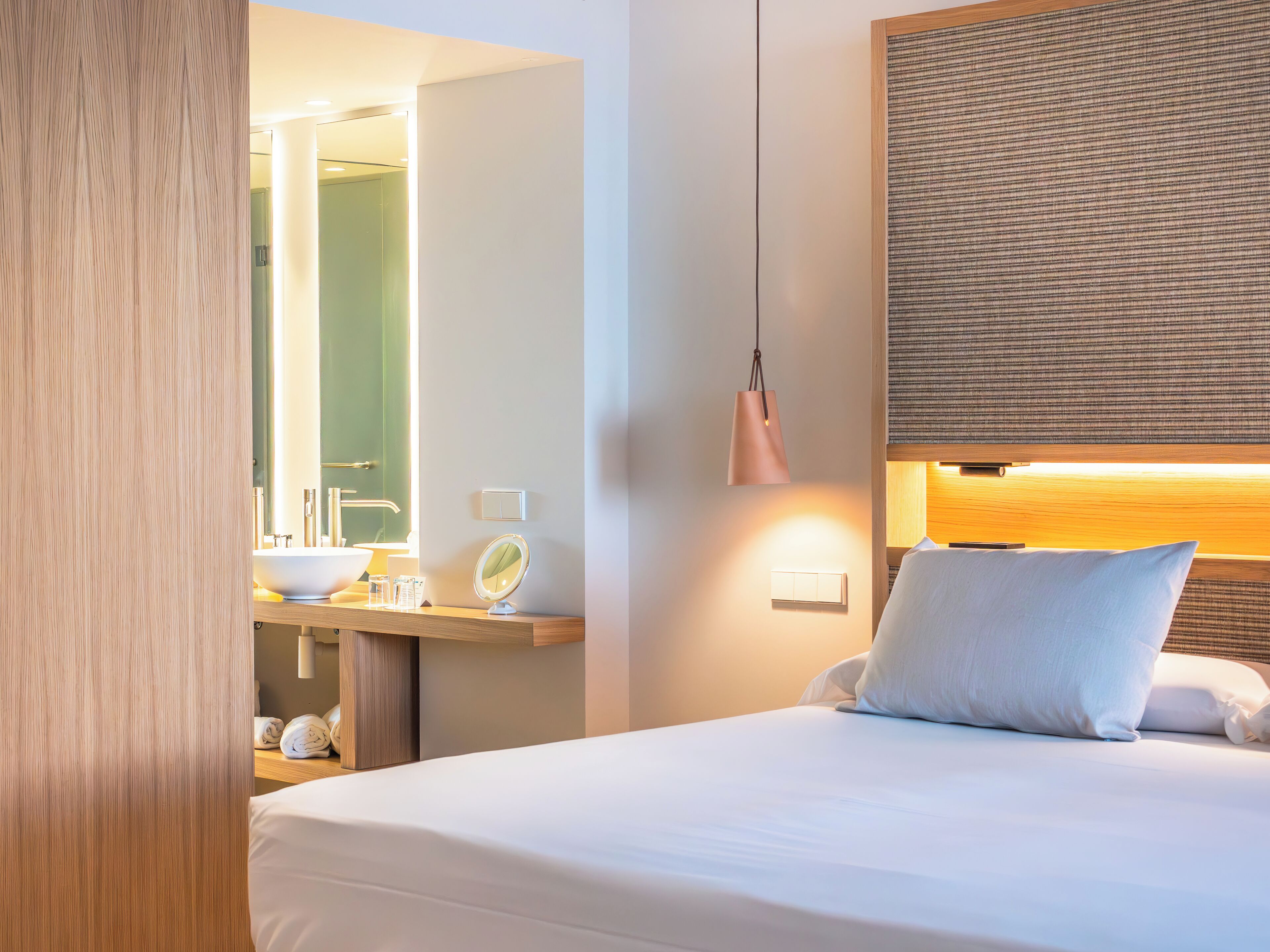 Habitación de hotel con cama, mesita de noche con espejo, lámpara y aplique de pared.