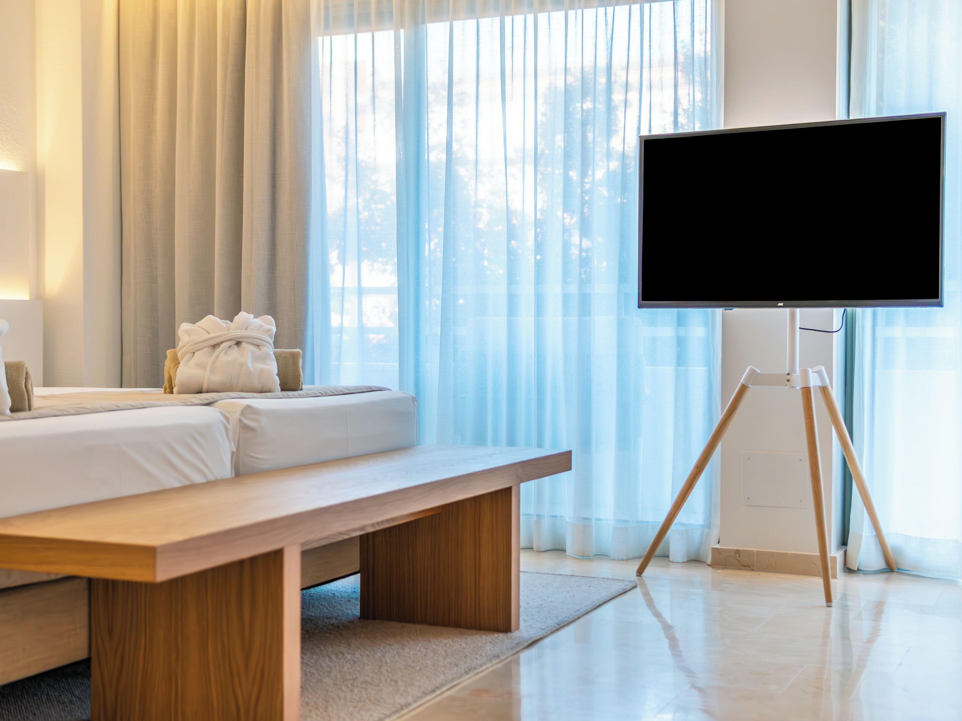 A hotel room with a bed, a wooden bench, a TV on a tripod, and sheer curtains.