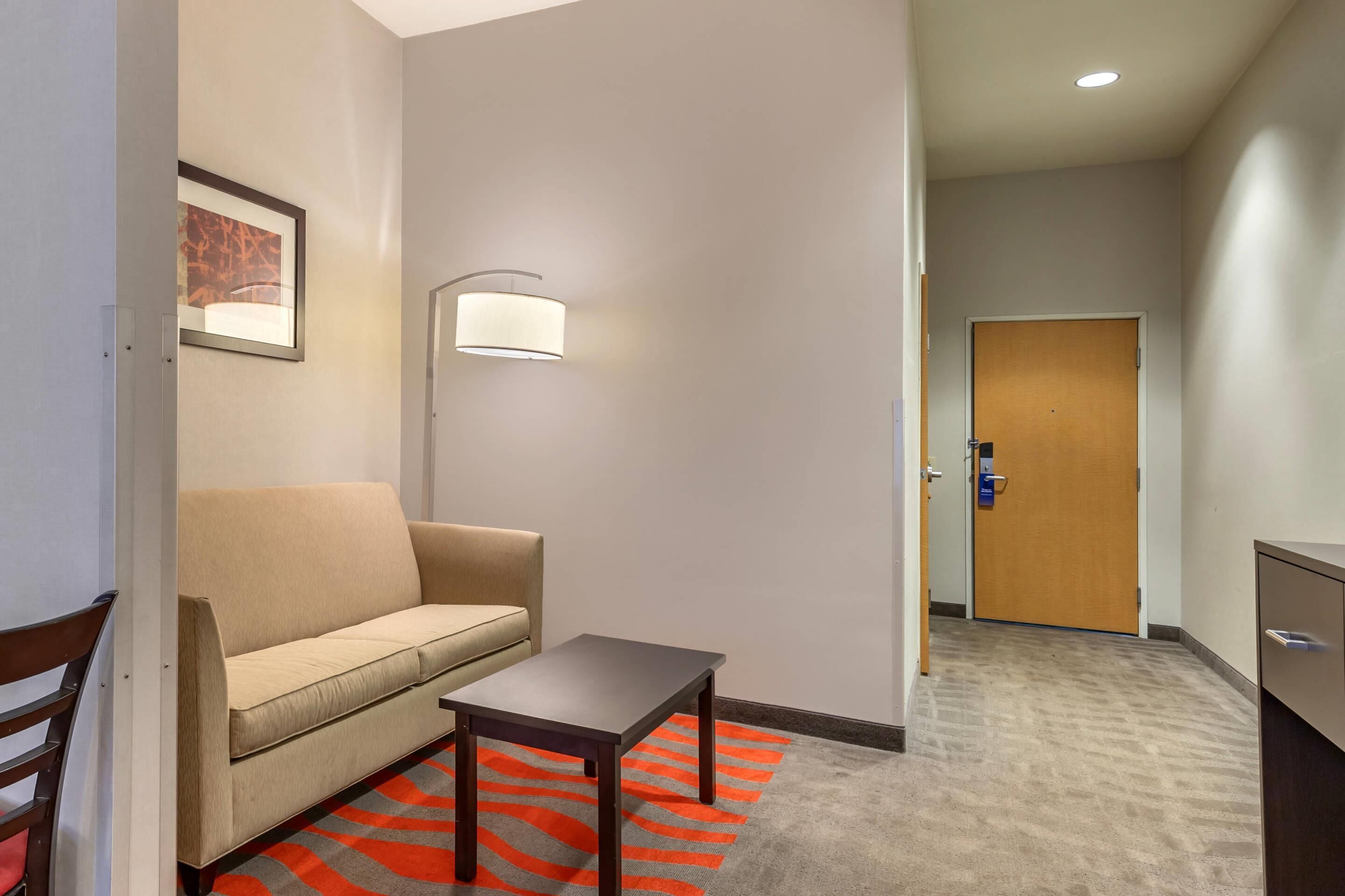 Habitació d'hotel amb sofà, taula petita, làmpada, porta i alfàbbra amb dibuix vermell i gris.