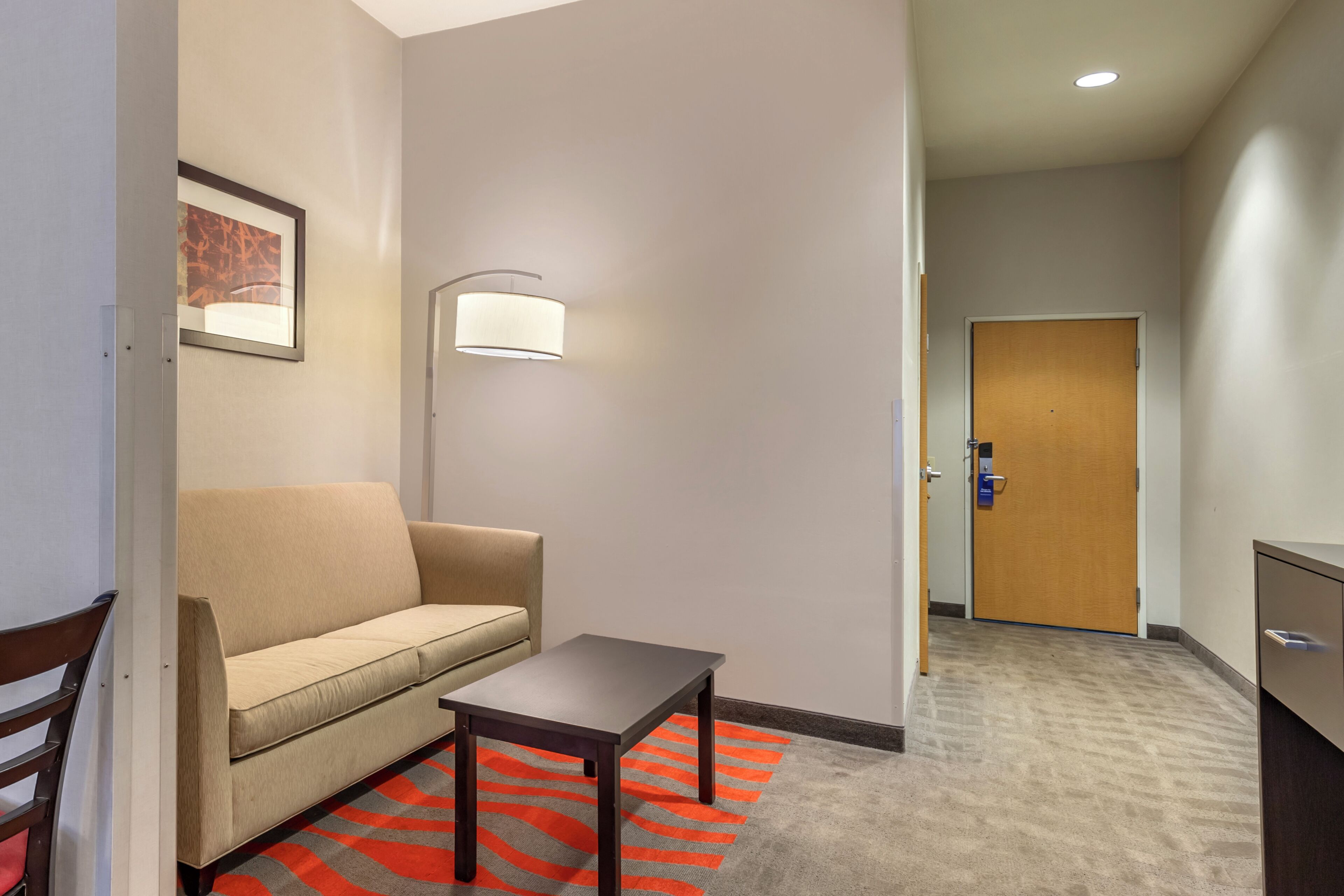A hotel room with a sofa, a small table, a lamp, a door, and a carpet with a red and gray pattern.