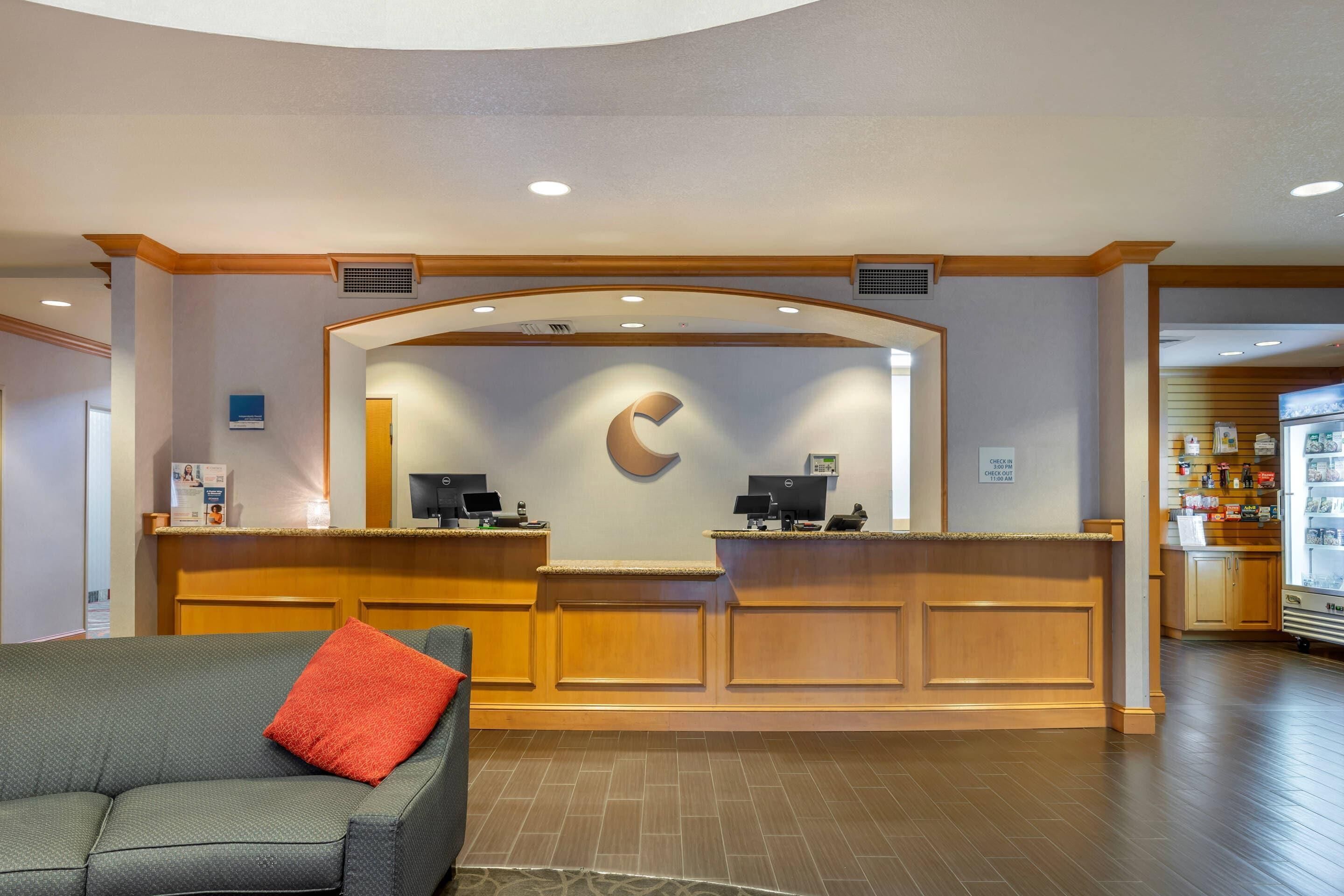 A reception area with a wooden reception desk, a large C-shaped logo on the wall, a grey sofa with a red pillow, and a kitchenette in the background.
