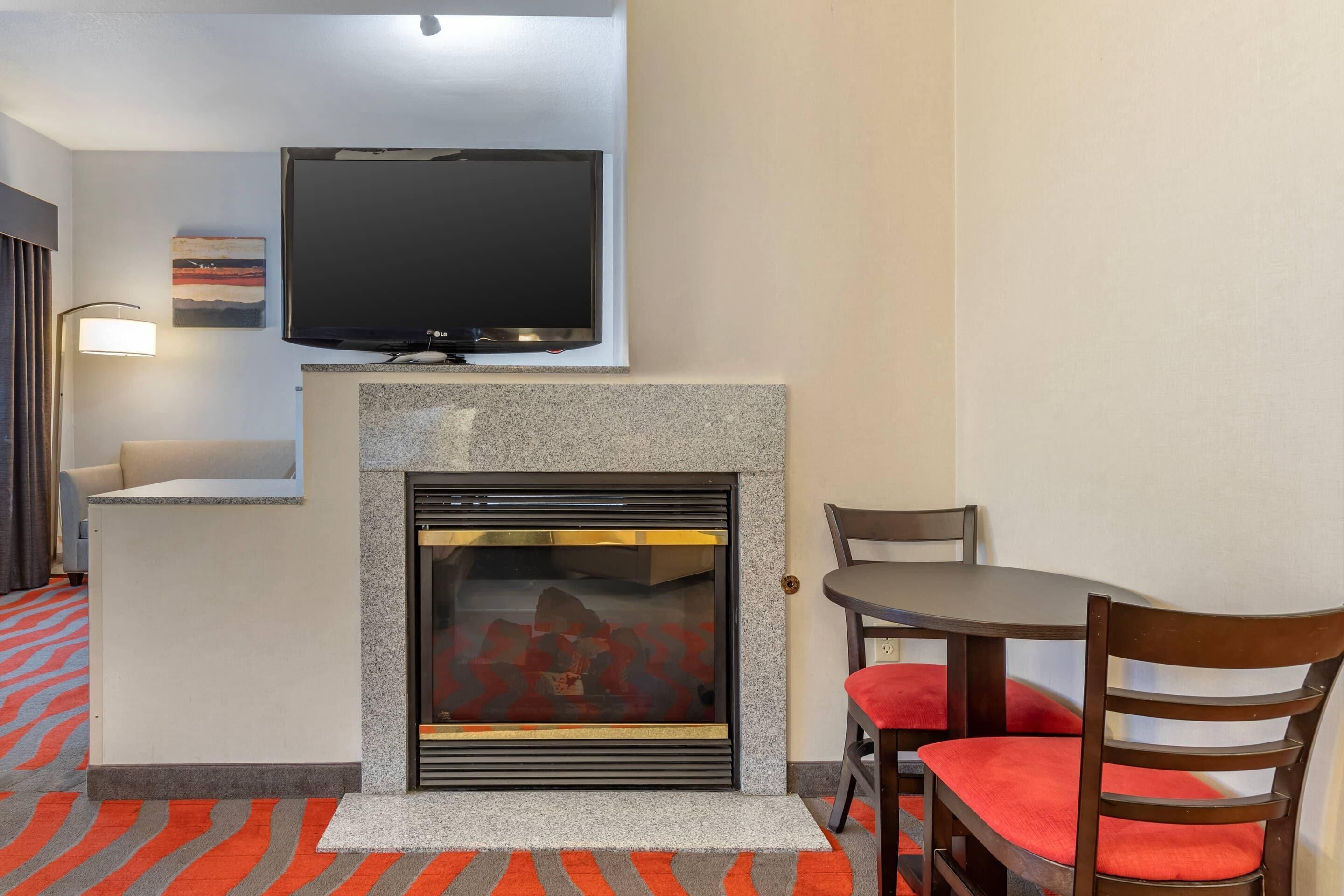 Een hotelkamer met een open haard, een televisie erboven, een ronde tafel met stoelen en een kamerbreed tapijt met dessin.
