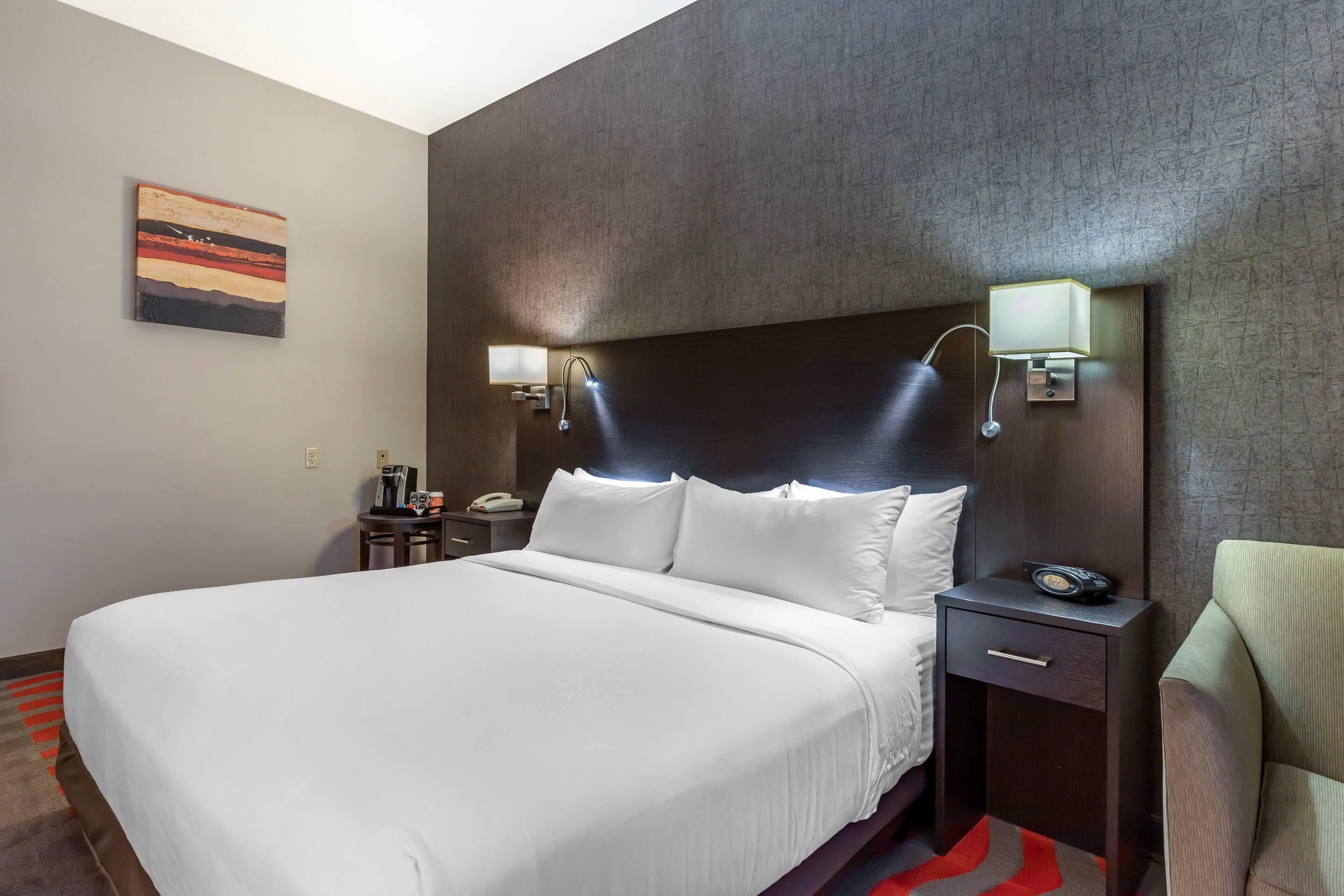 A hotel room with a large bed, a nightstand with a lamp, a chair, and a painting on the wall.