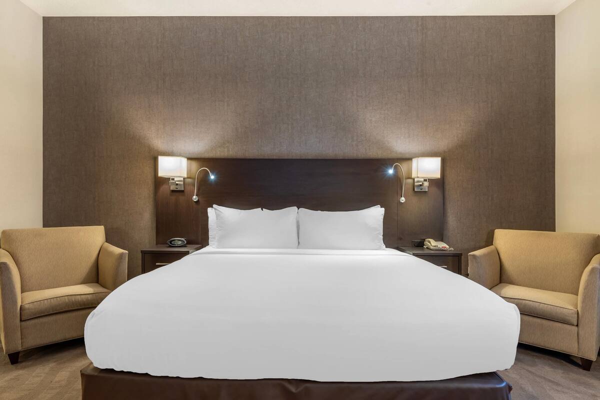 A hotel room with a large bed, two bedside tables, a headboard with wall-mounted lights, and two armchairs.