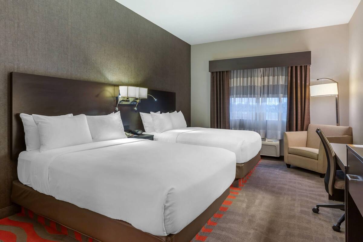A hotel room with two beds, a desk, a chair, a lamp, and a window with curtains.