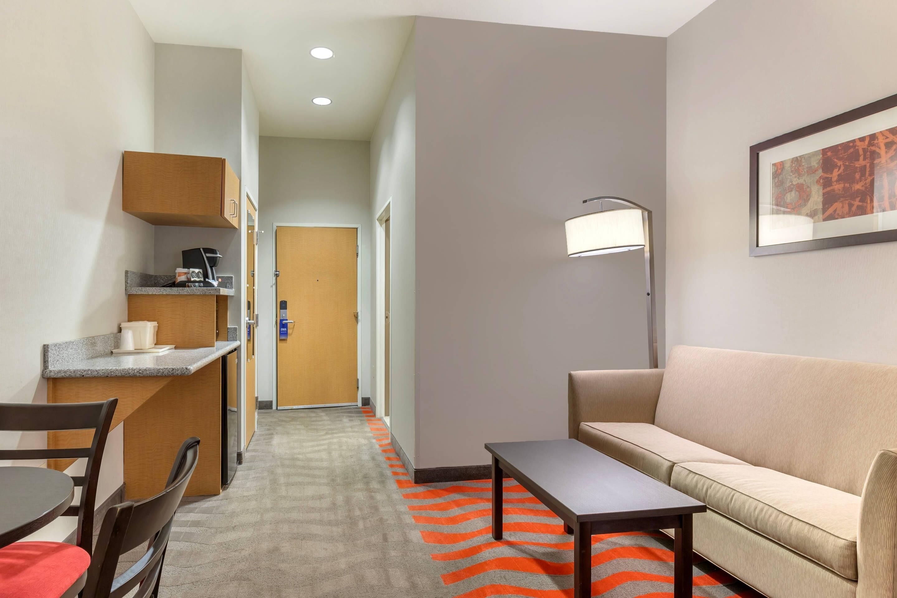 A hotel room with a kitchenette, a sofa, a table, and a hallway leading to a door.
