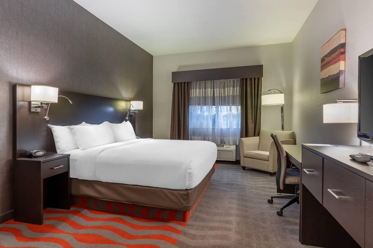 A hotel room with a bed, desk, chair, and a large window with curtains.