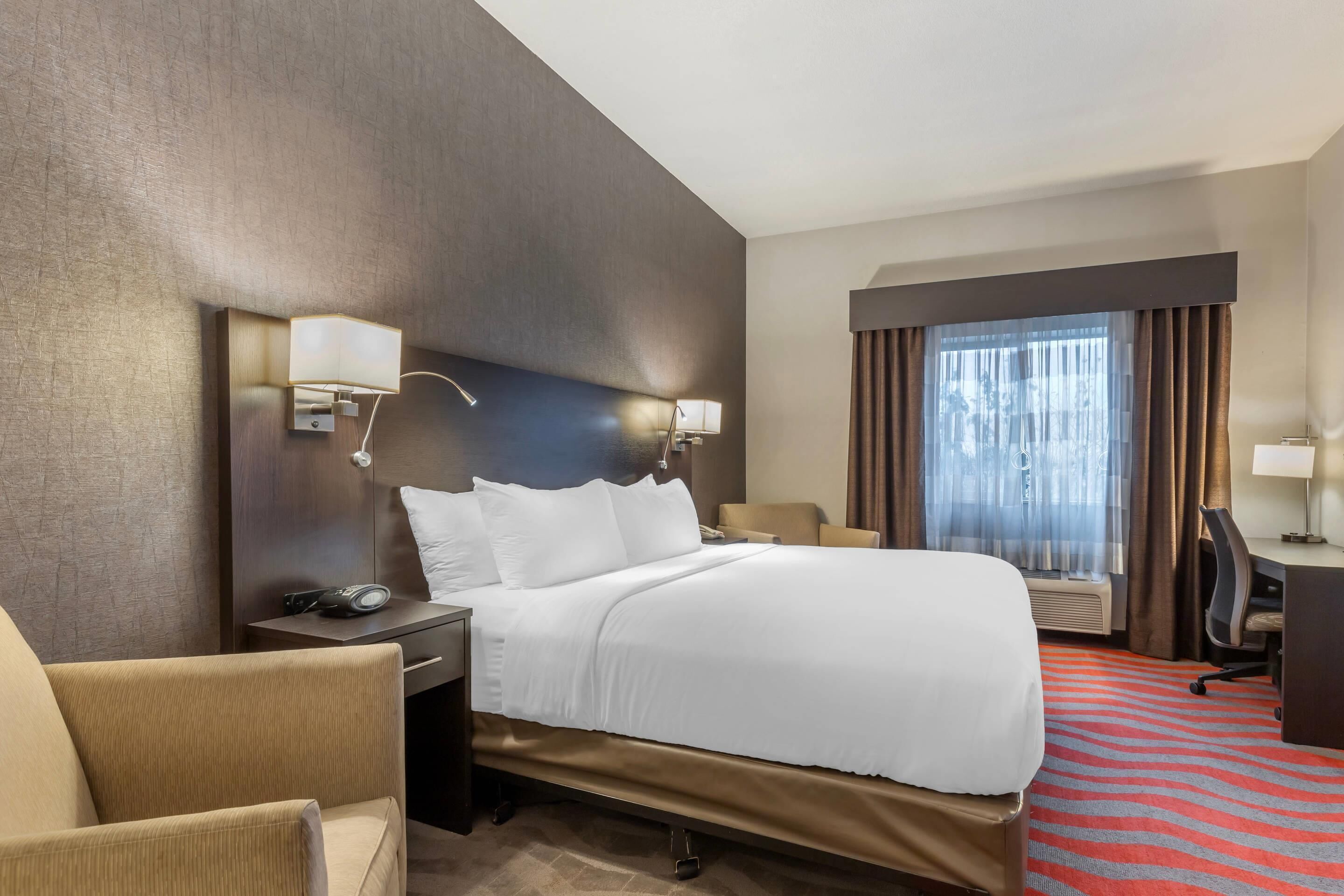 A hotel room with a bed, a desk, a chair, a lamp, and a window with curtains.