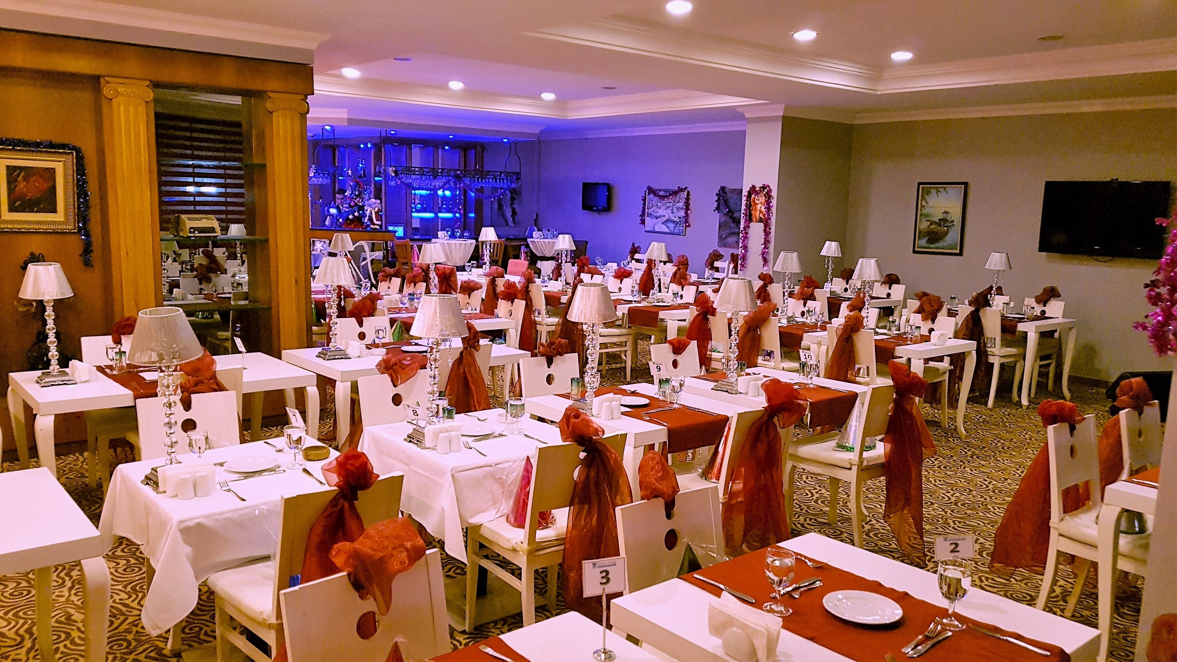 Un salón de banquetes con varias mesas dispuestas para comer, señalización con números y una zona de escenario al fondo.