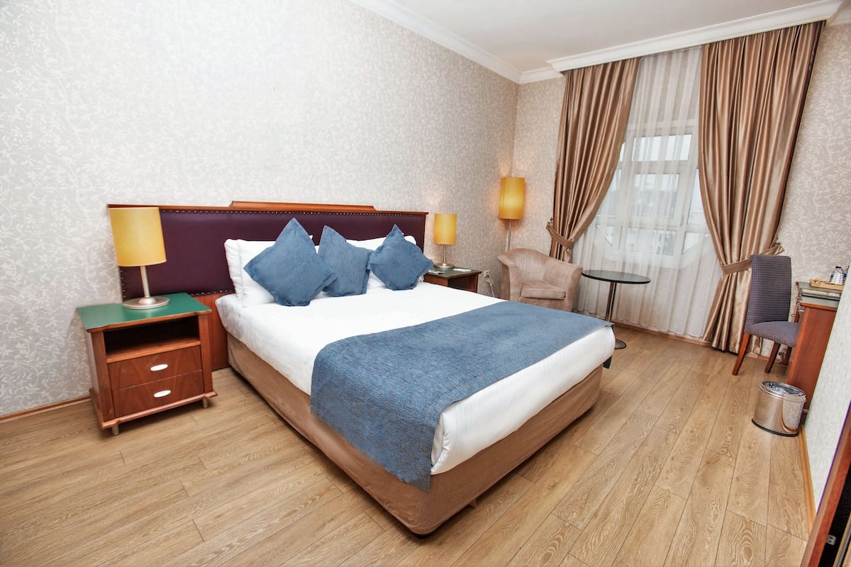 A hotel room with a large bed, two bedside tables, a chair, a small table, and a window with curtains.