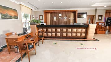 A hotel reception area with a wooden reception desk, a seating area with chairs, and a large painting on the wall.