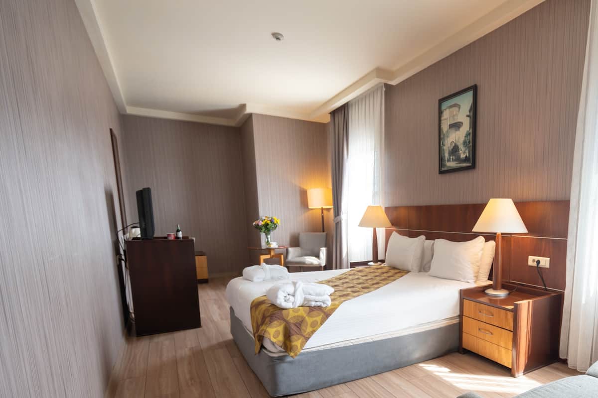 Quarto de hotel com uma cama grande, mesas de cabeceira com luminárias, uma escrivaninha, uma cadeira e um quadro na parede.