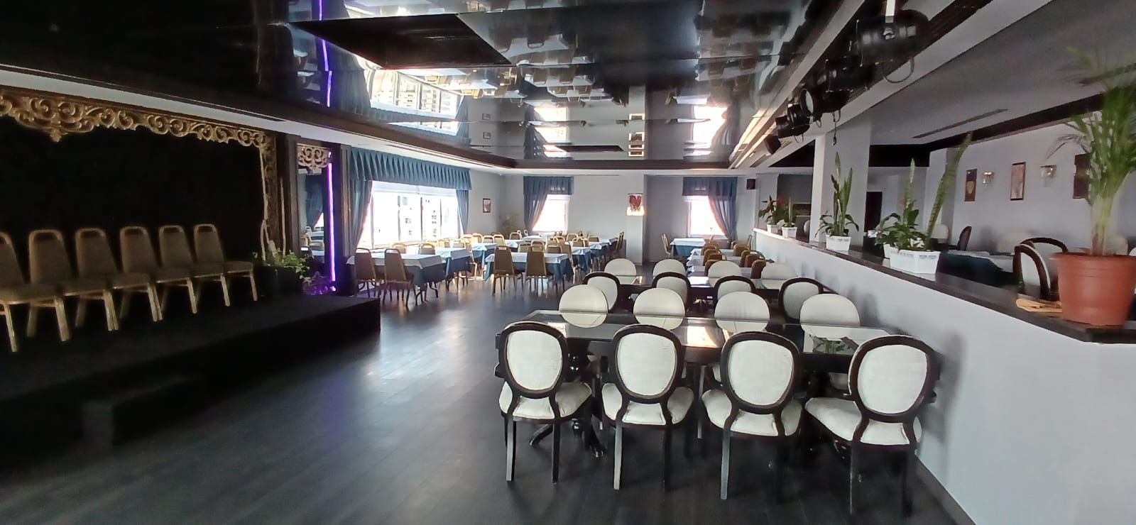 Un grand espace dédié à la salle à manger, avec de nombreuses tables et chaises, une scène sur la gauche et un long comptoir agrémenté de plantes en pot sur la droite.